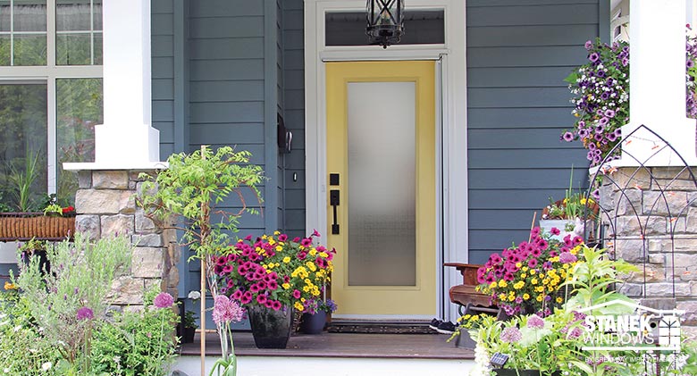 Yellow entry door with full privacy glass on blue-gray houe with stone and wood pillars, brightly colored potted flowers.

