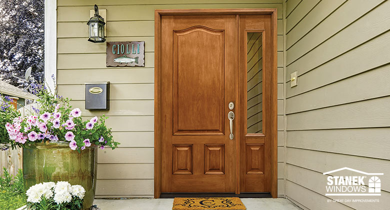 Cherry wood entry door with brass hardware and sidelite on a tan home with potted flowers on the front porch.
