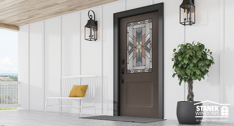 Art Deco glass inset on brown entry door with black hardware flanked by black lanterns on white farmhouse-style home.

