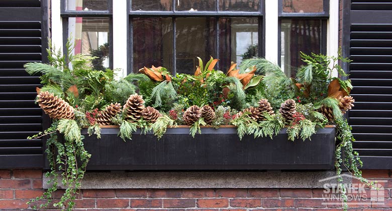 A black window box filled with pinecones and greenery on the outside of a window with black shutters.