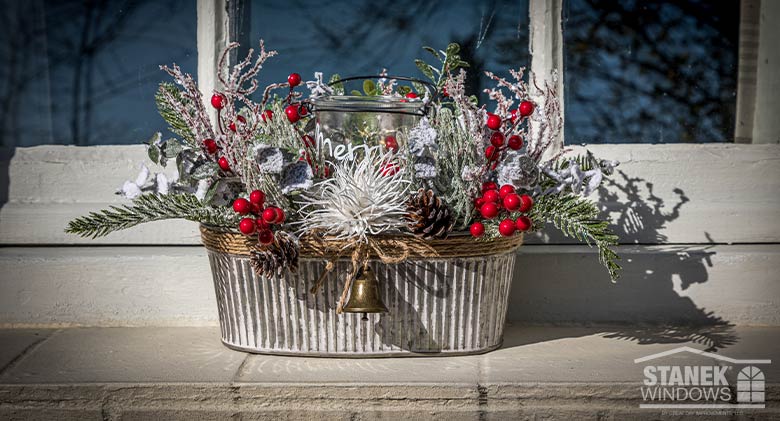 A tin window box on a window ledge with greenery, twigs, red berries, jute bow with bell and a glass candle holder in center.
