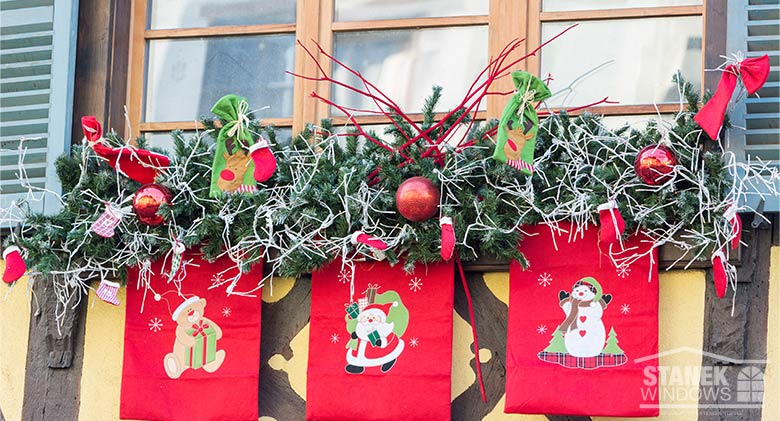 A window box decorated with greenery, ornaments, twigs, bows, and red banners hanging from the bottom with holiday characters.