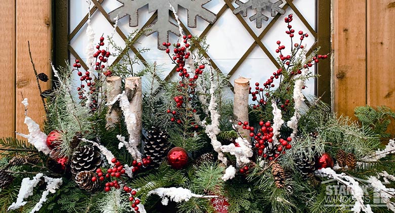 Holiday window box filled with branches, berries, pinecones, and evergreen branches. Snowflake decorations on the window.