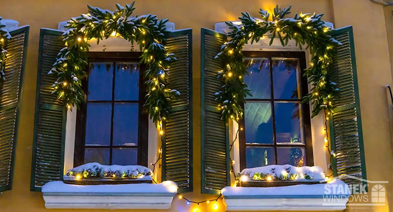At night, two windows with garland and window boxes of evergreens and white lights, covered with a layer of snow.